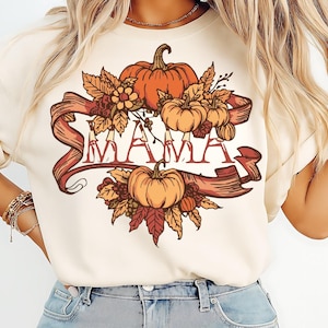 May include: Cream-colored t-shirt featuring a fall-themed design. The design includes pumpkins, leaves, and the word "MAMA" in a decorative font, all set against a ribbon banner. The overall color palette is warm, with shades of orange, brown, and red.