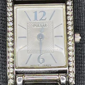 May include: Silver-toned rectangular watch with a white face. The watch face features the number 12 and 6, with the brand name "PULSAR" in the center. The watch is adorned with small, clear crystals around the face.
