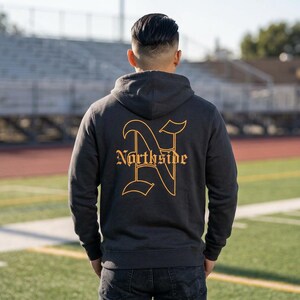 May include: Dark gray hoodie with a hood and long sleeves. The back of the hoodie features a large, stylized gold letter "N" with the word "Northside" beneath it in a gothic font. The person is wearing dark jeans.