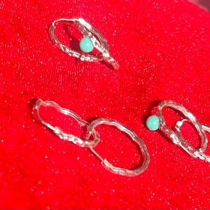 May include: Silver-toned earrings with a textured design and a small, round turquoise bead. The earrings are displayed on a bright red, glittery surface. The earrings have an open, oval shape.