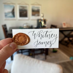 May include: A white card with the names "Whitney James" in elegant black calligraphy. A gold wax seal with a laurel wreath design is affixed to the card. The card is held up in front of a blurred interior background.