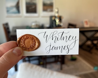 Può includere: Un biglietto bianco con i nomi "Whitney James" in elegante calligrafia nera. Un sigillo di cera dorata con un disegno a corona d'alloro è apposto sul biglietto. Il biglietto è tenuto davanti a uno sfondo interno sfocato.