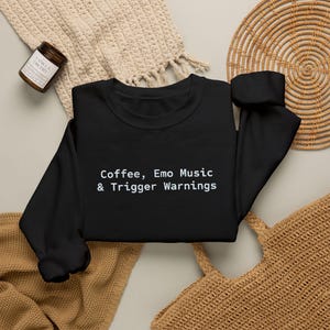 May include: A black crewneck sweatshirt featuring white text printed on the chest that reads, "Coffee, Emo Music & Trigger Warnings." The garment has long sleeves, a ribbed collar, and ribbed cuffs, presented in a clean, straightforward design.