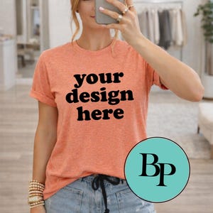 May include: Orange short-sleeved t-shirt with the text "your design here" in black. The model is wearing light blue jeans. A turquoise circle with the letters "Bp" is in the lower right corner.