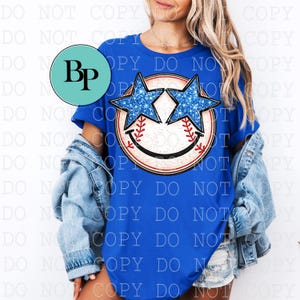 May include: Royal blue t-shirt featuring a baseball-themed graphic. The graphic includes a baseball with a smiling face and star-shaped sunglasses with blue glitter. The shirt is worn with a denim jacket.
