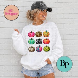 May include: A white long-sleeve sweatshirt featuring a grid of nine pumpkins with various colorful patterns, including leopard print, polka dots, and bright neon hues. The sweatshirt is paired with light blue denim shorts and a dark gray baseball cap.
