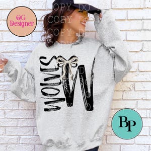 May include: A light gray crewneck sweatshirt featuring a black graphic print of the word "WOLVES" with a decorative, glittery silver bow placed between the letters. The sweatshirt has long sleeves and a relaxed fit, made of a soft, heathered fabric.