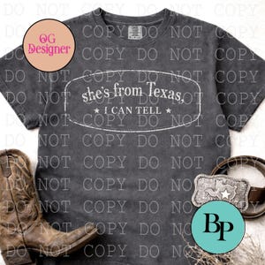 May include: Dark gray t-shirt with the text "she's from Texas, I can tell" in a white oval. The image includes a brown leather belt with a Texas-shaped buckle and a pair of brown cowboy boots.
