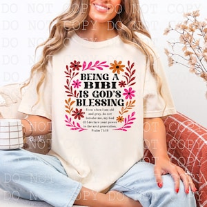 May include: Cream-colored t-shirt with the text "BEING A BIBI IS GOD'S BLESSING" in black, surrounded by a floral design in shades of pink, orange, and brown. The shirt also includes a Bible verse.
