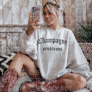 Könnte beinhalten: Ein weißes Sweatshirt mit dem Text "Champagne problems" in schwarzer gotischer Schrift. Das Sweatshirt hat lange Ärmel und einen Rundhalsausschnitt. Das Bild zeigt auch braune Cowboy-Stiefel und ein Mobiltelefon.