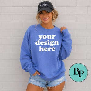 May include: A blue crewneck sweatshirt featuring the white text your design here on the front. The garment has long sleeves and a ribbed collar, cuffs, and hem. A black baseball cap with the white text Nashville Est. 1779 is also included.