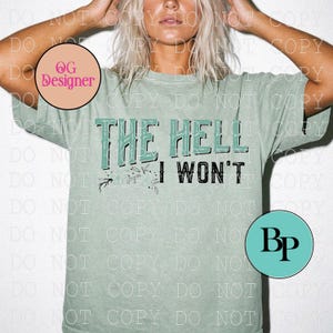 May include: A sage green short-sleeve t-shirt featuring the text THE HELL I WON'T printed in a distressed black and teal font. The design includes small floral line art accents below the text, set against a solid, muted green fabric background.
