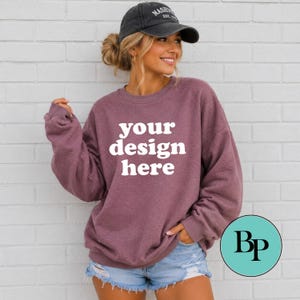 Może przedstawiać: Wrzosowa bluza z długim rękawem i okrągłym dekoltem z białym napisem your design here z przodu, zestawiona z jasnoniebieskimi szortami dżinsowymi i ciemnoszarą czapką z daszkiem z białym haftowanym napisem Nashville Est. 1779.