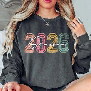 A woman with long blonde hair is sitting on a bed, wearing a black sweatshirt with the text "2026" printed on it.