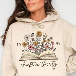 May include: Beige hoodie with a front graphic of an open book with flowers and butterflies. The text "chapter thirty" is below the book. The numbers "19" and "95" are on either side of the floral design.
