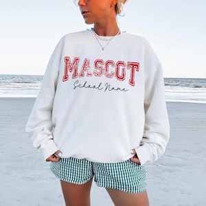 May include: A white sweatshirt with the word "MASCOT" in red block letters and "School Name" in a cursive font. The sweatshirt is worn with green and white striped shorts. The background is a beach scene.