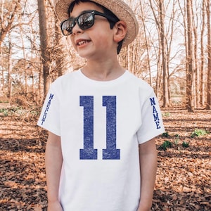 A young boy wearing a white t-shirt with the number 11 and sunglasses stands in a wooded area.