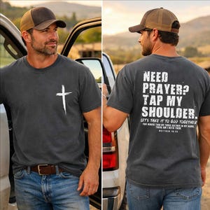 May include: A charcoal gray t-shirt with a white cross graphic on the front and the text "NEED PRAYER? TAP MY SHOULDER" on the back. The shirt also features the text "LET'S TAKE IT TO GOD TOGETHER" and "MATTHEW 18:20". The person is wearing a brown and tan baseball cap.