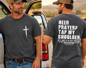 May include: A charcoal gray t-shirt with a white cross graphic on the front and the text "NEED PRAYER? TAP MY SHOULDER" on the back. The shirt also features the text "LET'S TAKE IT TO GOD TOGETHER" and "MATTHEW 18:20". The person is wearing a brown and tan baseball cap.