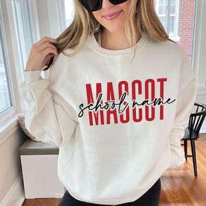 May include: Cream-colored sweatshirt with the words "MASCOT SCHOOL NAME" in red block letters and black cursive script. The sweatshirt has long sleeves and a crew neck. The person in the image is wearing sunglasses.