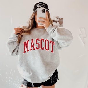 May include: A light gray sweatshirt with the word "MASCOT" in red block letters. The sweatshirt is paired with a black baseball cap and black shorts. The cap has white text.