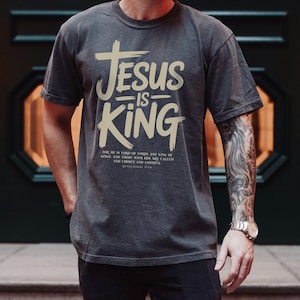 A man wearing a gray t-shirt with the text "JESUS IS KING" printed on it, standing in front of a dark background.