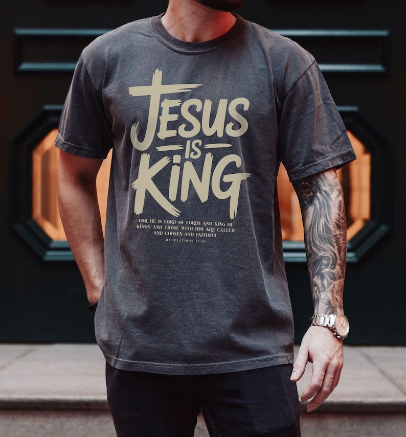 A man wearing a gray t-shirt with the text "JESUS IS KING" printed on it, standing in front of a dark background.
