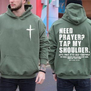 May include: An olive green hoodie with a white cross on the front and the text "NEED PRAYER? TAP MY SHOULDER" on the back. The hoodie also has a front pocket and a drawstring hood. The text "LET'S TAKE IT TO GOD TOGETHER" is also printed on the back.