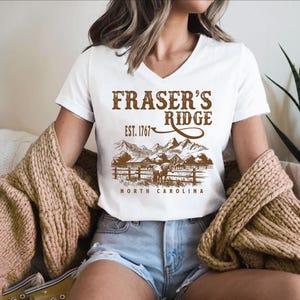 A woman wearing a white t-shirt with a graphic design of a mountain range and the text "Fraser's Ridge" printed on it. She is also wearing denim shorts and a beige sweater.