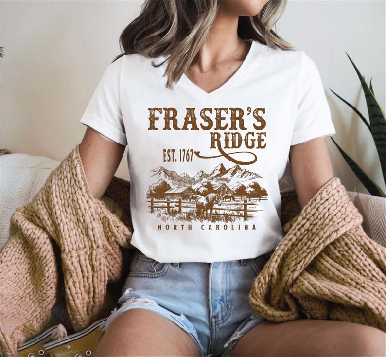 A woman wearing a white t-shirt with a graphic design of a mountain range and the text "Fraser's Ridge" printed on it. She is also wearing denim shorts and a beige sweater.