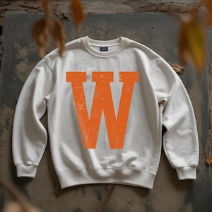 May include: Cream-colored sweatshirt with a large, distressed orange letter "W" across the front. The sweatshirt has a crew neck and long sleeves. The background is a textured, neutral surface with some leaves.