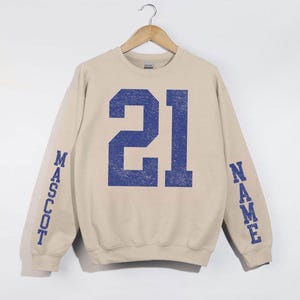 A beige sweatshirt with the number 21 in blue printed on it, hanging on a wooden hanger.