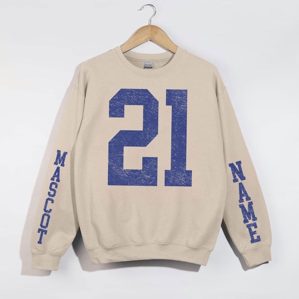 May include: Beige sweatshirt with blue number 21 on the front. The sleeves have the words "MASCOT" and "NAME" printed in blue. The sweatshirt is hanging on a wooden hanger.