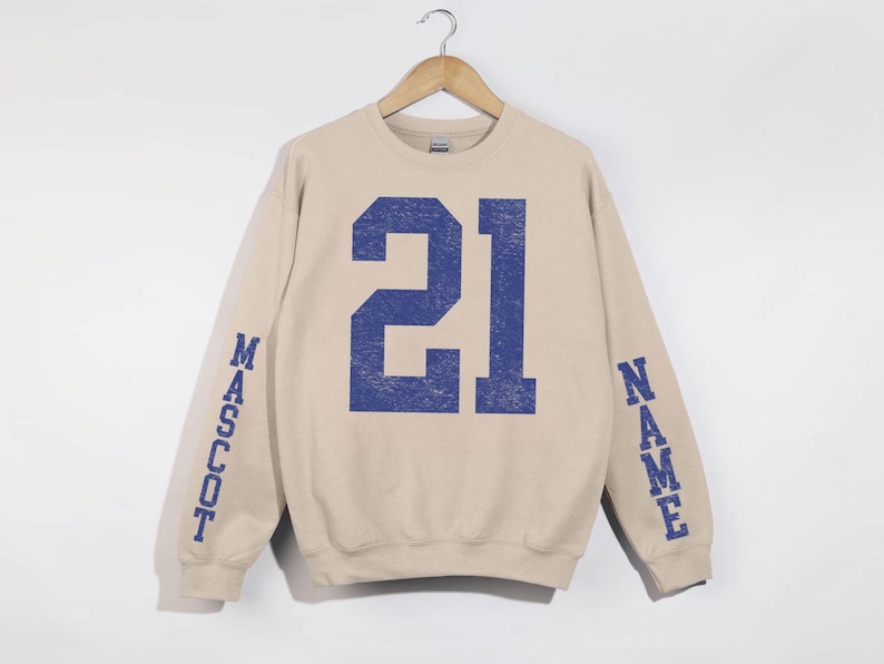 A beige sweatshirt with the number 21 in blue printed on it, hanging on a wooden hanger.