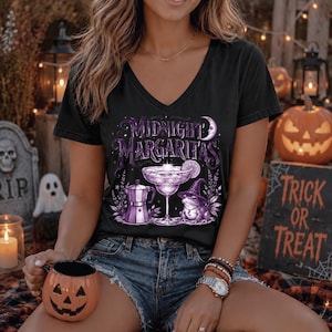 May include: A black V-neck t-shirt featuring a purple graphic print with the text Midnight Margaritas, a cocktail glass, a moka pot, a crescent moon, and a frog wearing a witch hat. The design is centered on the front of the short-sleeved shirt.