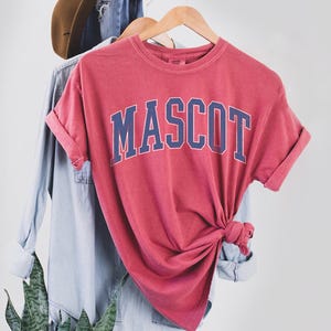 May include: A red t-shirt with the word "MASCOT" in blue block letters. The shirt is knotted at the bottom and hangs on a wooden hanger with a denim shirt and a brown hat in the background.