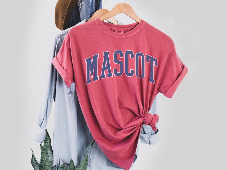 A red t-shirt with the word "MASCOT" printed on it, hanging on a clothing hanger.