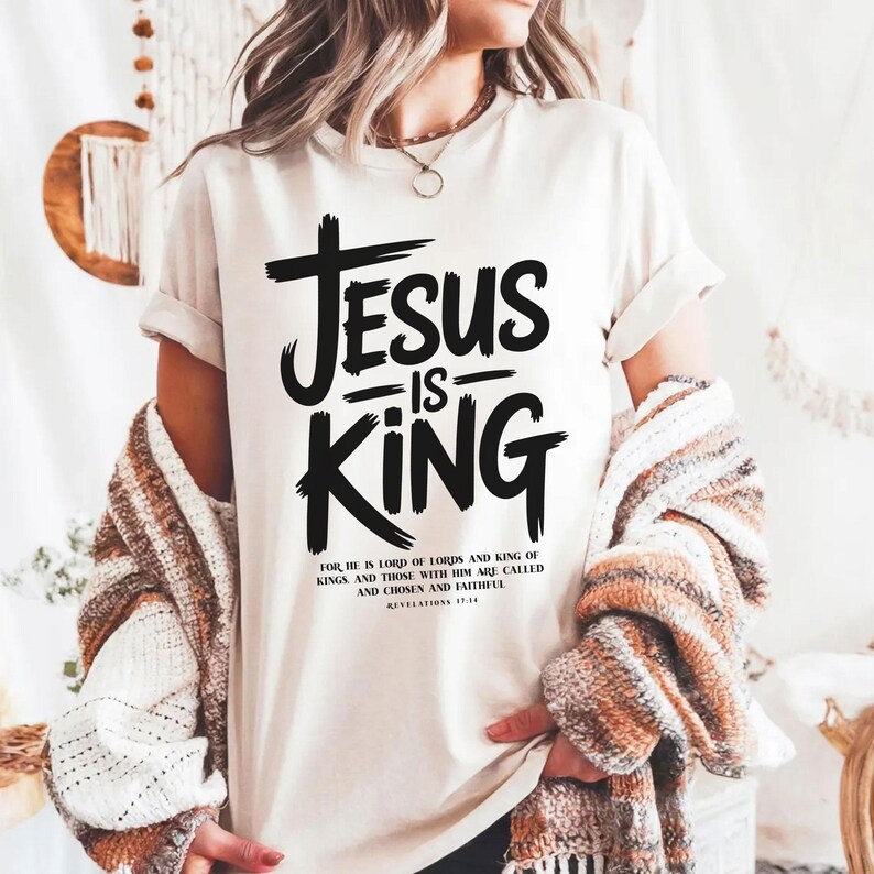A person wearing a white t-shirt with the text "JESUS IS KING" printed on it, along with a Bible verse reference.