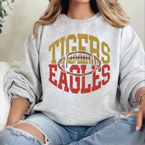 May include: A light gray sweatshirt with the words "TIGERS" and "EAGLES" in gold and red, respectively, surrounding a football graphic. The sweatshirt is a casual style.