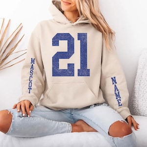 A woman wearing a beige hoodie with the number 21 and the word "MASSOT" printed on it, sitting on a couch.