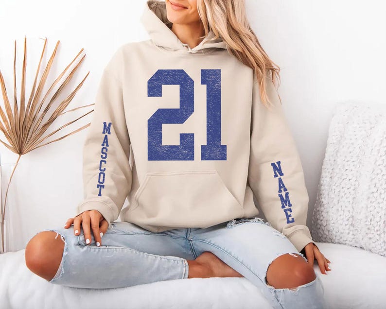 A woman wearing a beige hoodie with the number 21 and the word "MASSOT" printed on it, sitting on a couch.