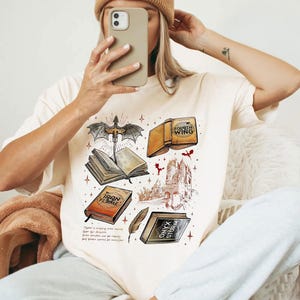 A person is sitting down and taking a selfie with a phone, wearing a white t-shirt with a graphic design featuring various books and a dragon.