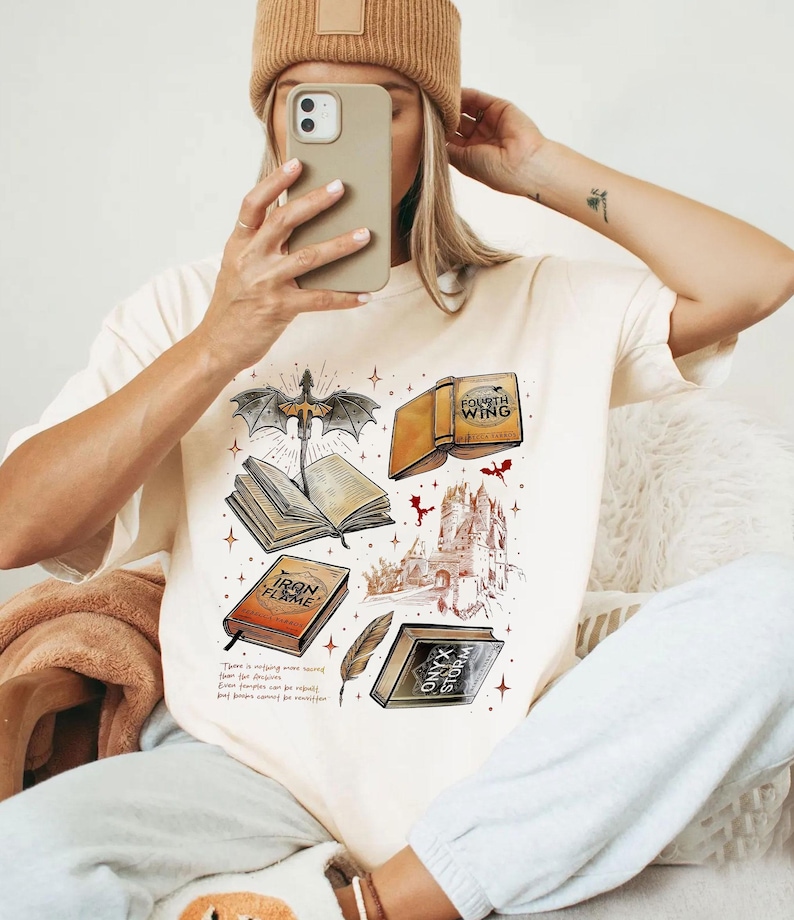 A person is sitting down and taking a selfie with a phone, wearing a white t-shirt with a graphic design featuring various books and a dragon.