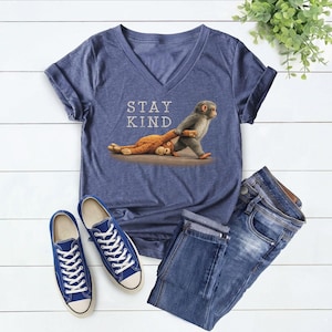 May include: A blue V-neck t-shirt with the words "STAY KIND" and an illustration of a monkey pulling a stuffed animal. The image also includes blue sneakers and denim jeans.