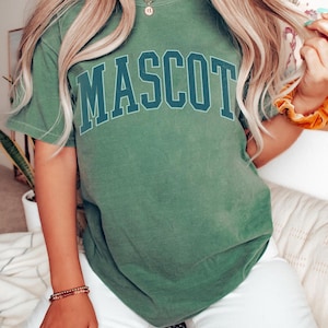 A person wearing a green t-shirt with the word "Mascot" printed on it, sitting on a bed with white sheets.