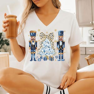 May include: White V-neck t-shirt featuring a Christmas design. The design includes two blue and gold nutcrackers flanking a blue and white Christmas tree with a black and white bow. Gold star accents and gift boxes complete the festive look.
