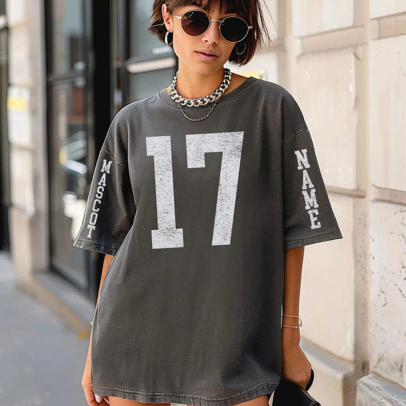 A woman wearing a grey t-shirt with the number 17 and the word "MASSOT" printed on it, standing in front of a building.