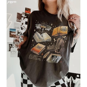 A person wearing a black t-shirt with a graphic design featuring various books and a bat, along with some text.