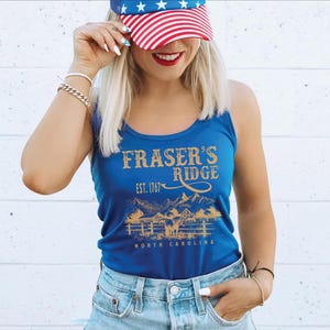 A woman wearing a blue tank top with the text "Fraser's Ridge" and a red, white, and blue American flag on her head, smiling and posing against a white brick wall.