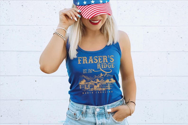 A woman wearing a blue tank top with the text "Fraser's Ridge" and a red, white, and blue American flag on her head, smiling and posing against a white brick wall.